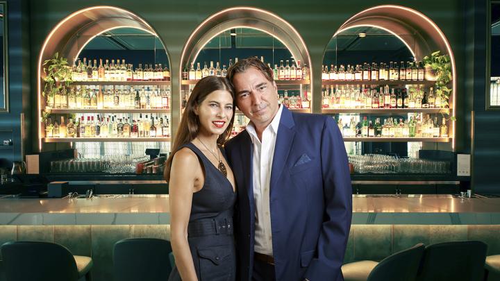 David Manica and his wife Noelle standing in front of a moody lit bar with bottles in the background.