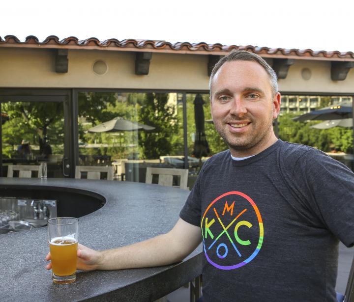 Man sitting at a bar with a beer, wearing a KCMO Pride shirt