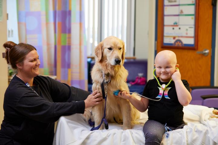 Child holding up a stethoscope to a dog, with a nurse smiling