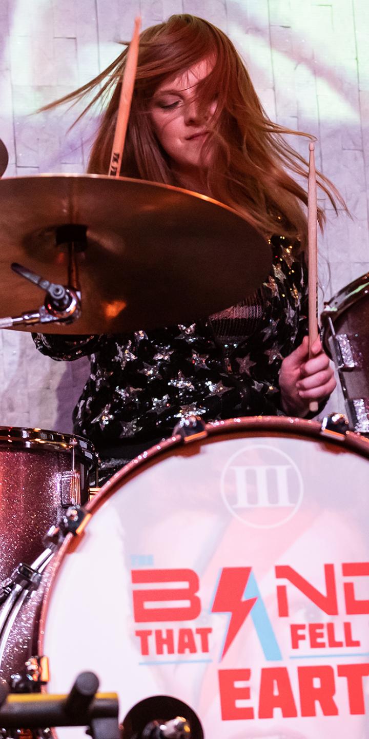 Woman playing a drumset with a logo saying Band That Fell to Earth